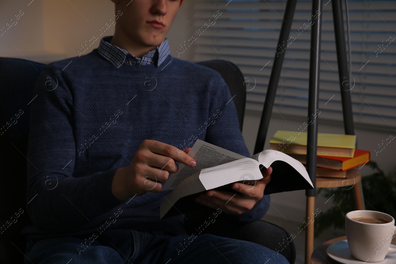 Photo of Man with cup of drink reading book in room at night, closeup