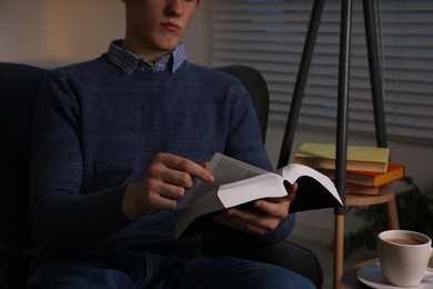 Man with cup of drink reading book in room at night, closeup