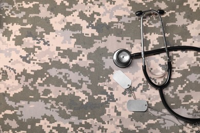 Photo of Stethoscope and military ID tags on camouflage fabric, flat lay. Space for text