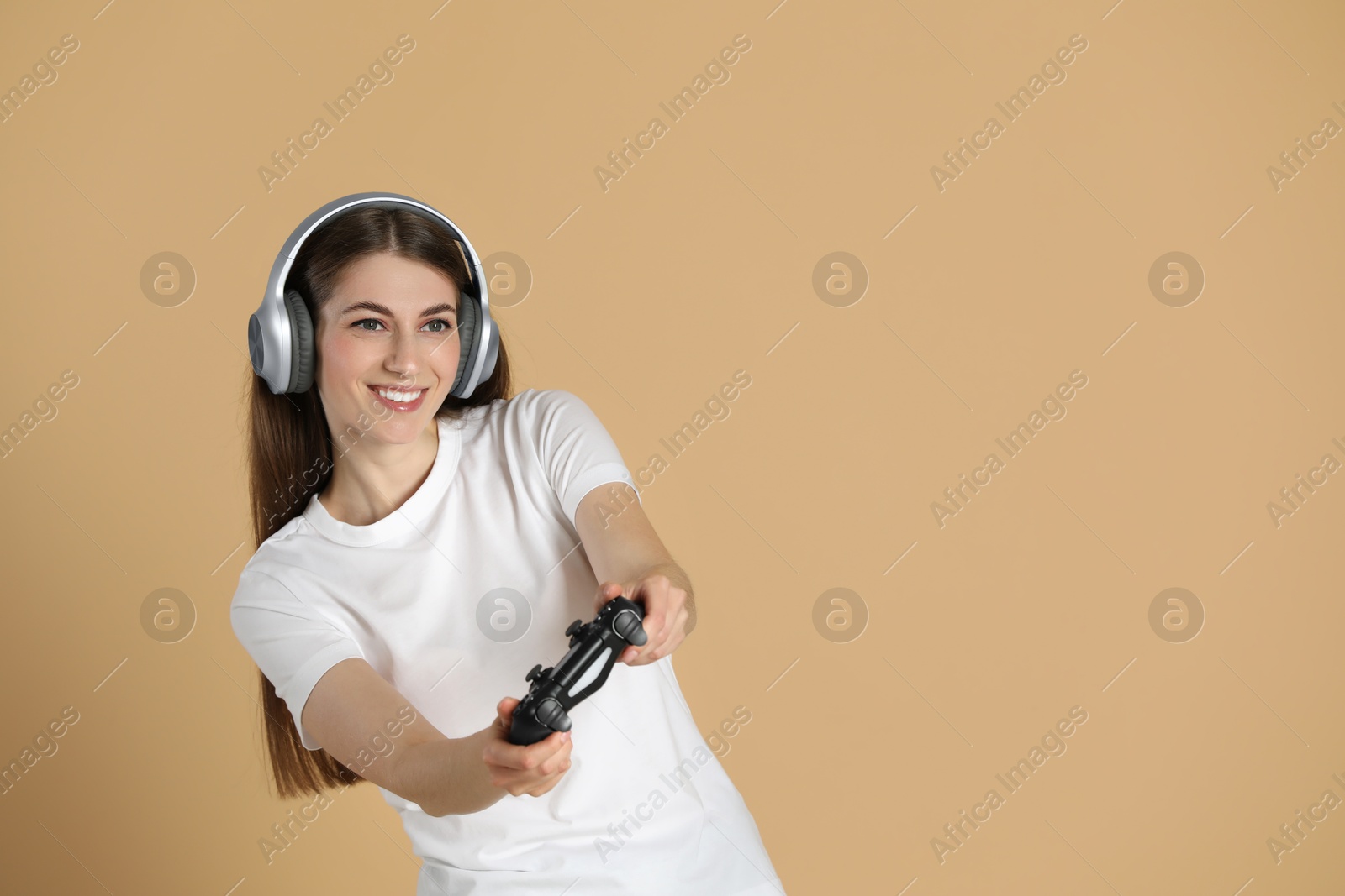 Photo of Happy woman in headphones playing video games with controller on beige background, space for text