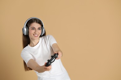 Happy woman in headphones playing video games with controller on beige background, space for text