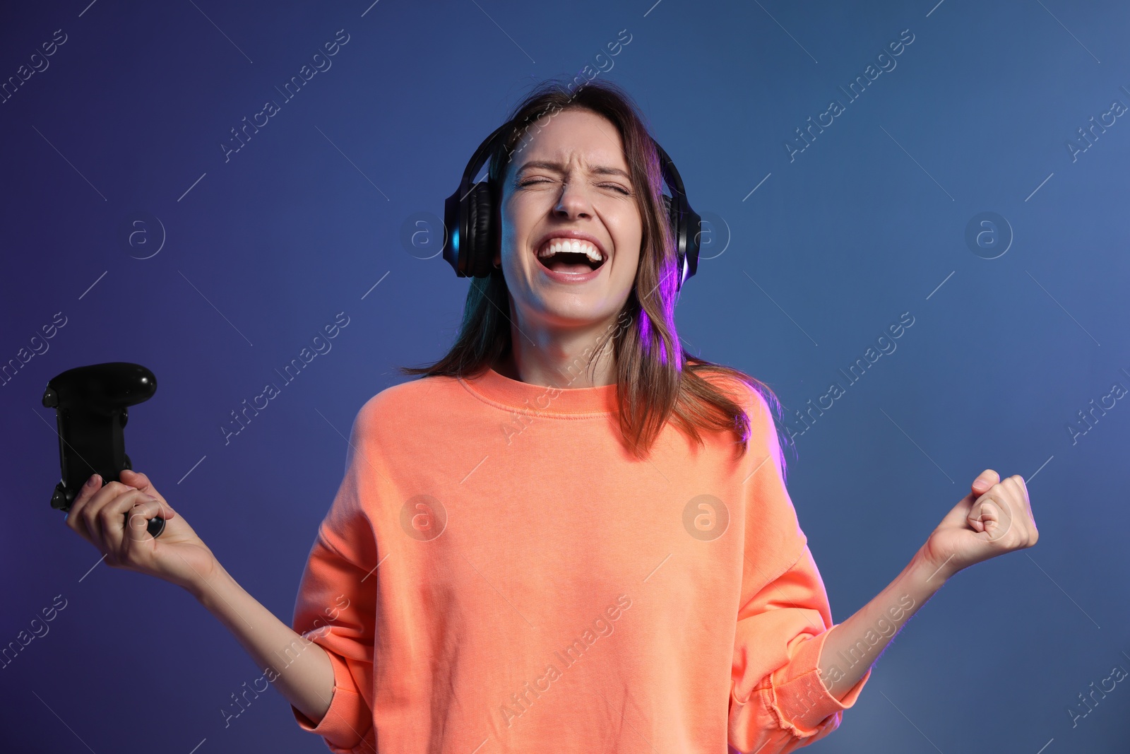 Photo of Emotional woman in headphones with game controller on dark blue background