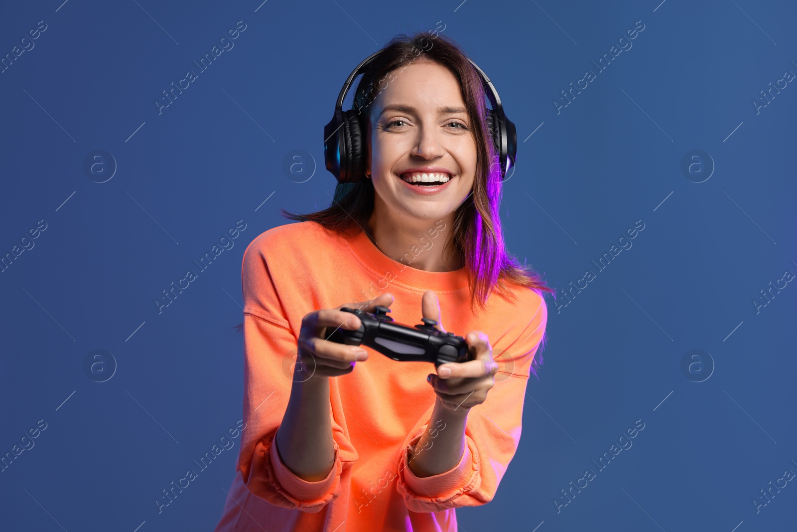 Photo of Happy woman in headphones playing video game with controller on dark blue background