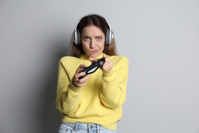 Emotional woman in headphones playing video game with controller on light grey background