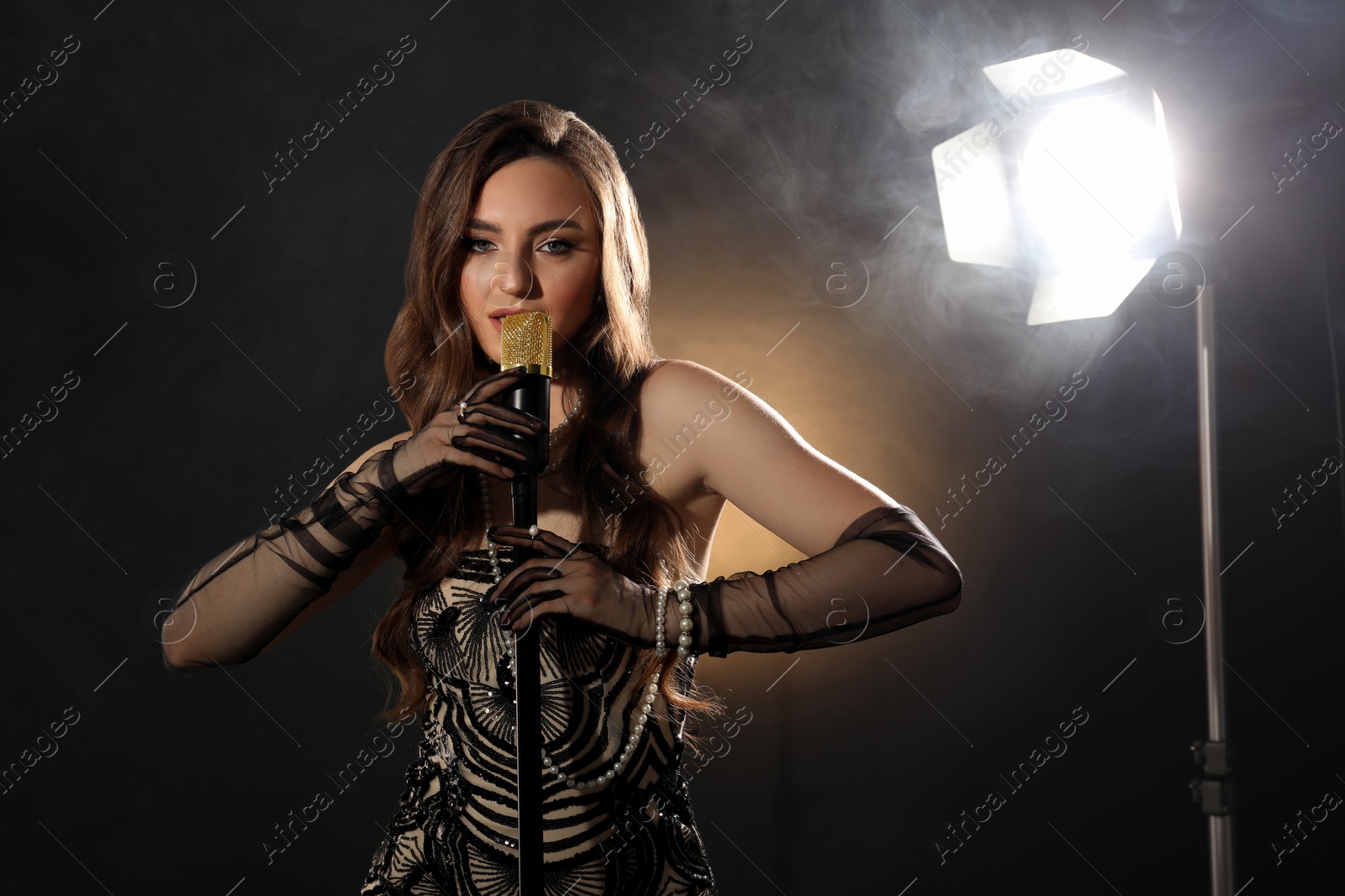 Photo of Beautiful young woman with microphone singing on dark background with smoke