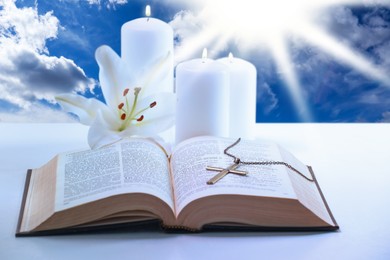 Holy Bible, cross, burning candles and lily flower on table against blue sky. Religion of Christianity