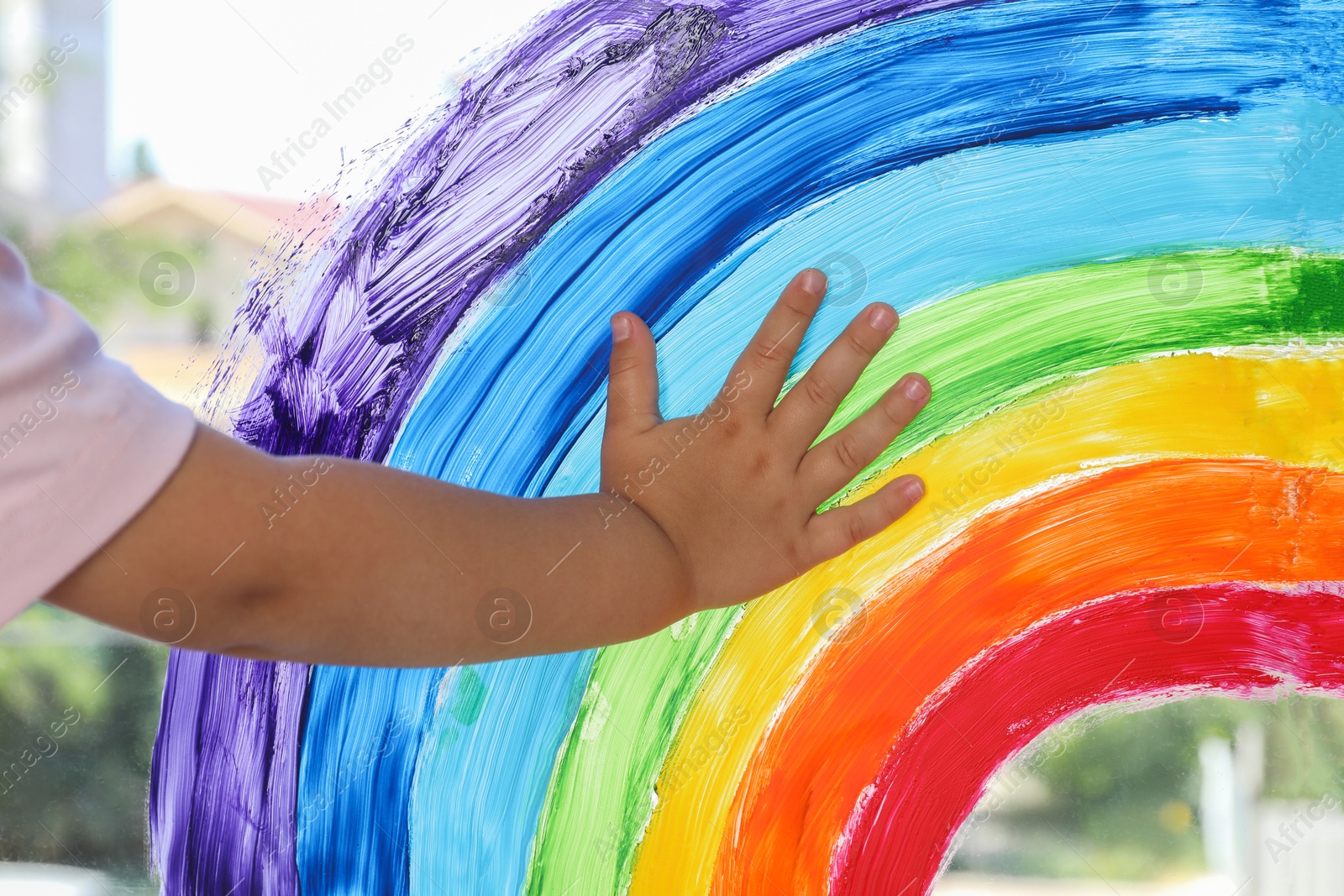 Photo of Little girl touching picture of rainbow on window indoors, closeup