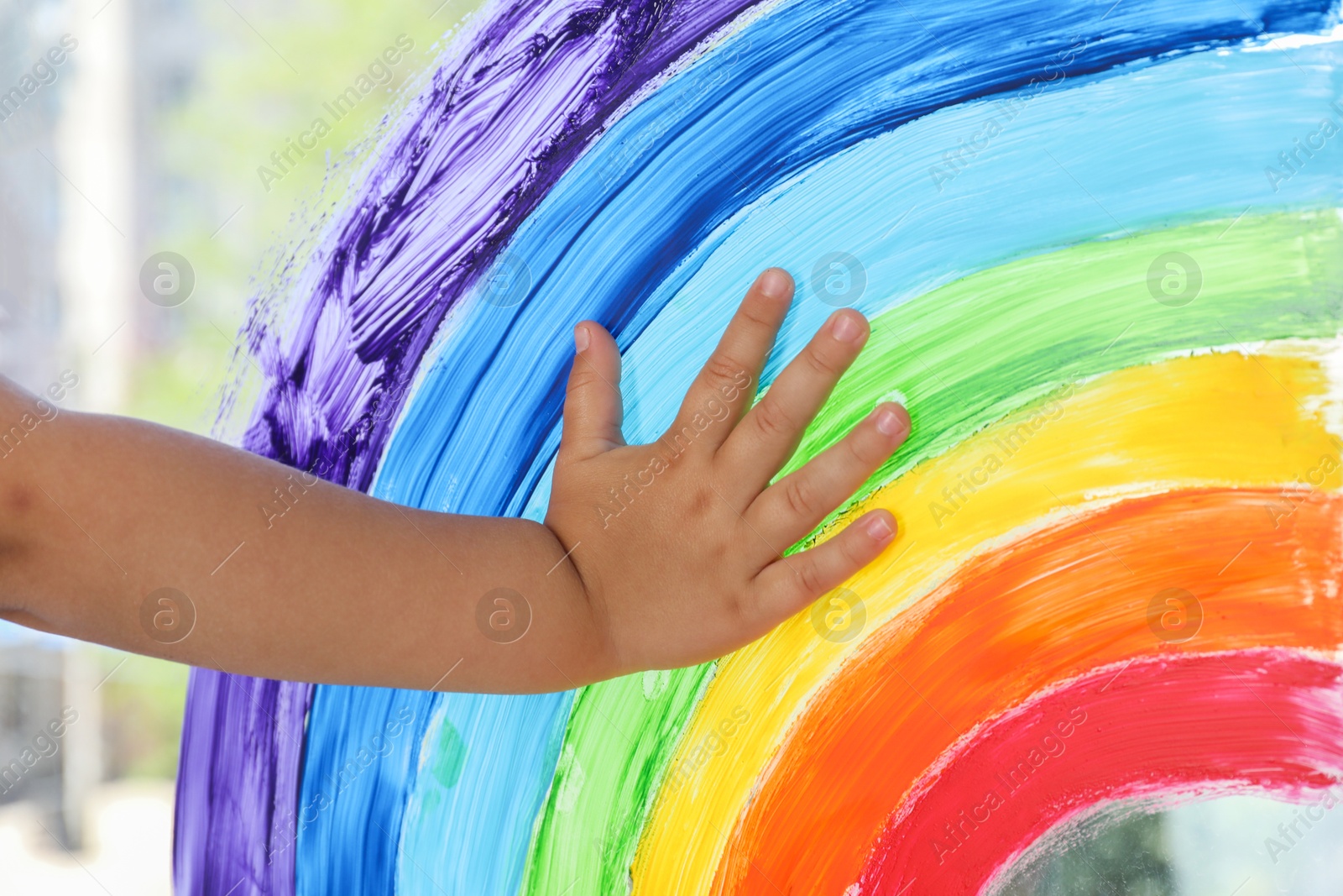 Photo of Little girl touching picture of rainbow on window indoors, closeup