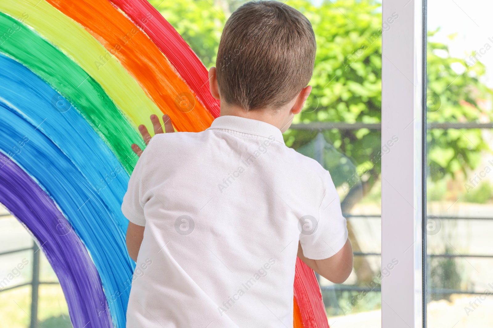 Photo of Little boy touching picture of rainbow on window indoors, back view