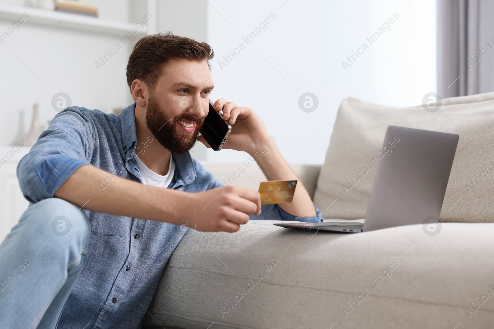 Photo of Online banking. Happy young man with credit card talking by smartphone at home