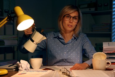Photo of Overwhelmed woman pouring coffee past cup at workplace in office at night