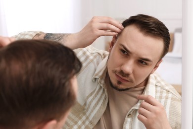 Photo of Baldness concept. Sad man with receding hairline looking at mirror indoors