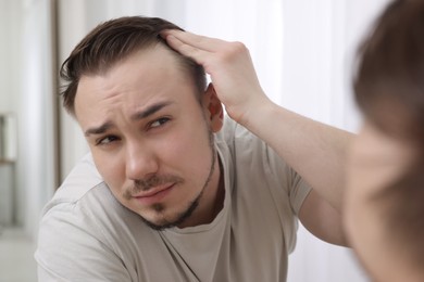 Baldness concept. Sad man with receding hairline looking at mirror indoors