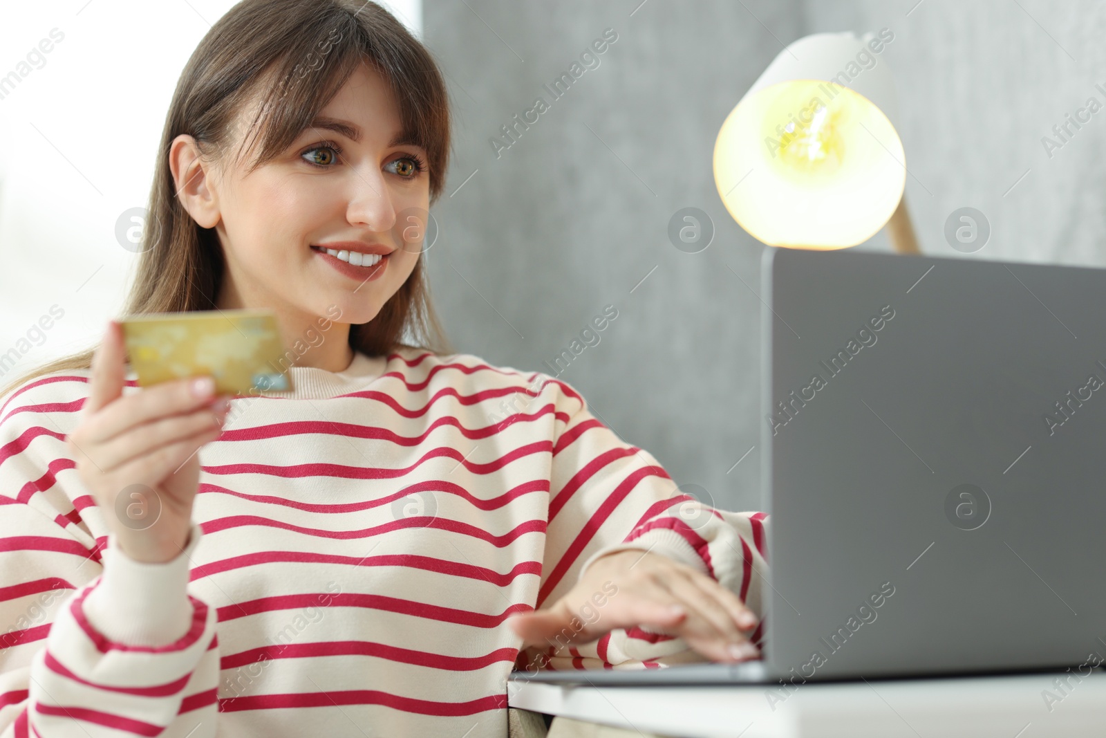 Photo of Online banking. Smiling woman with credit card and laptop paying purchase at table indoors