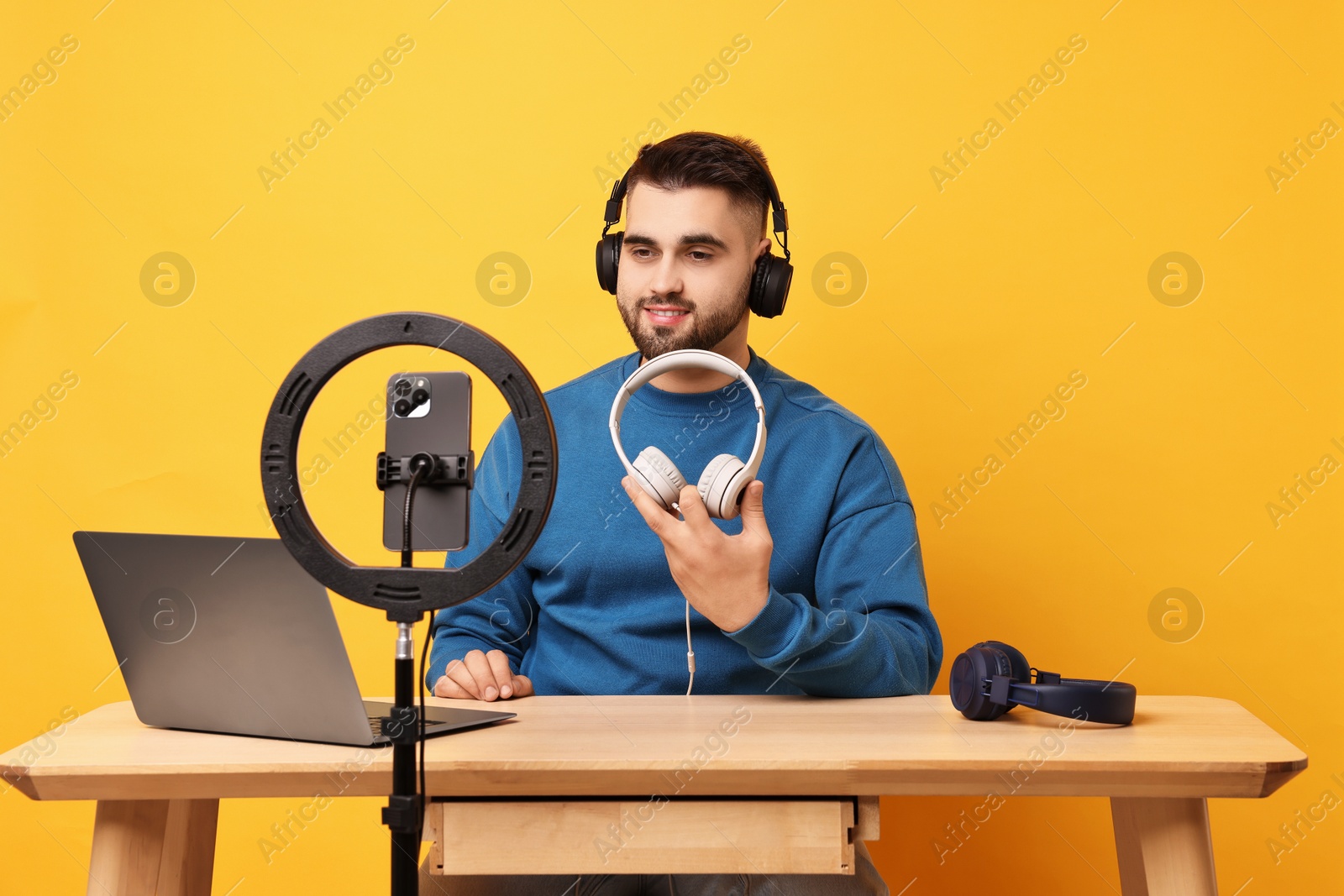 Photo of Technology blogger reviewing headphones and recording video with smartphone and ring lamp at wooden table on orange background