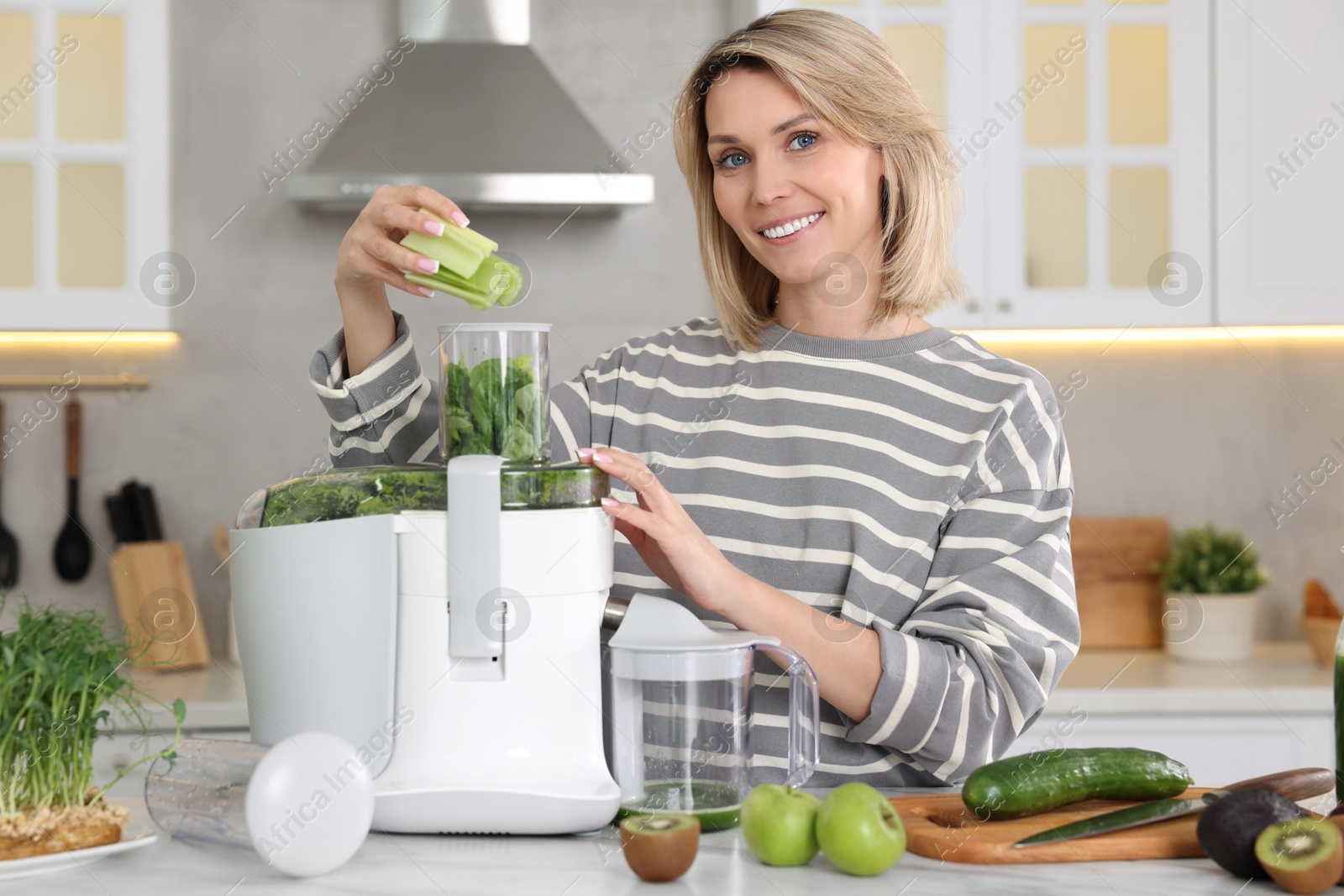 Photo of Smiling woman putting fresh celery into juicer at white marble table in kitchen