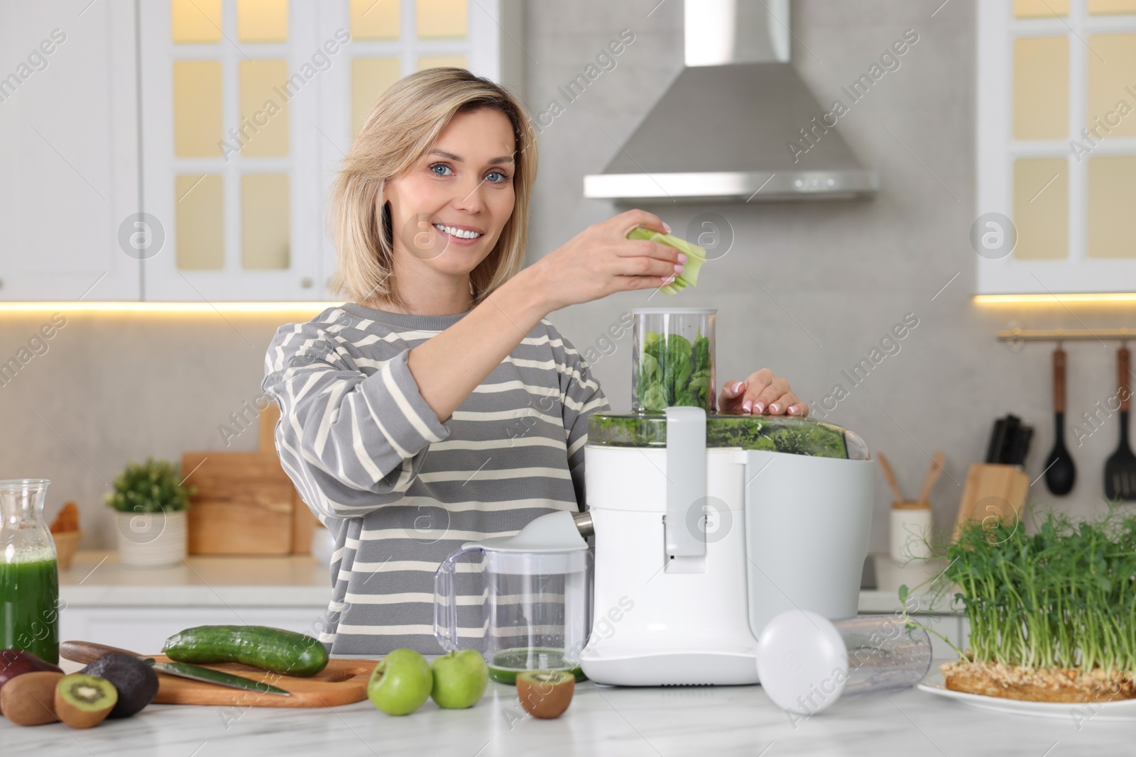 Photo of Smiling woman putting fresh celery into juicer at white marble table in kitchen