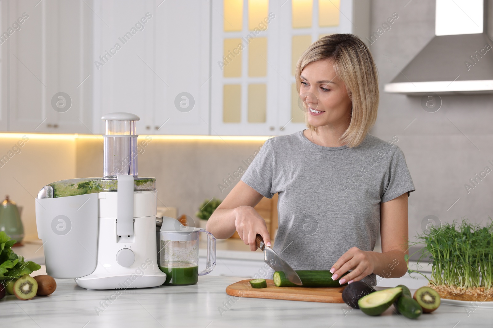 Photo of Juicer and fresh products on white marble table. Smiling woman cutting cucumber in kitchen