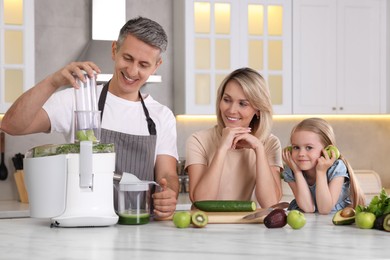 Photo of Happy family with juicer and fresh products making drink at white marble table in kitchen