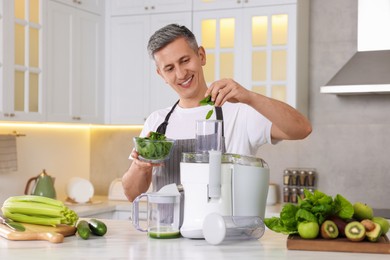 Photo of Smiling man putting fresh basil into juicer at white marble table in kitchen