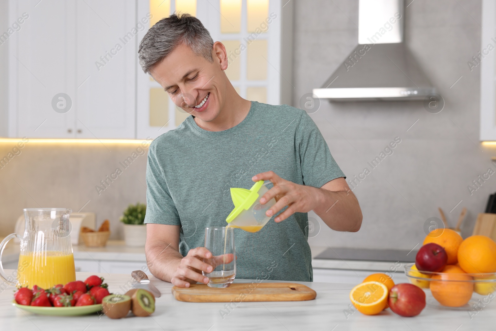 Photo of Smiling man pouring fresh orange juice into glass at white marble table in kitchen