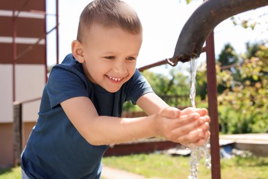 Photo of Water scarcity. Cute little boy drawing water with hands from tap outdoors