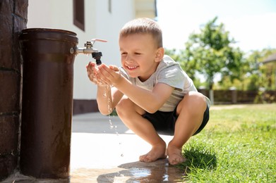 Photo of Water scarcity. Cute little boy drinking water from tap outdoors, space for text