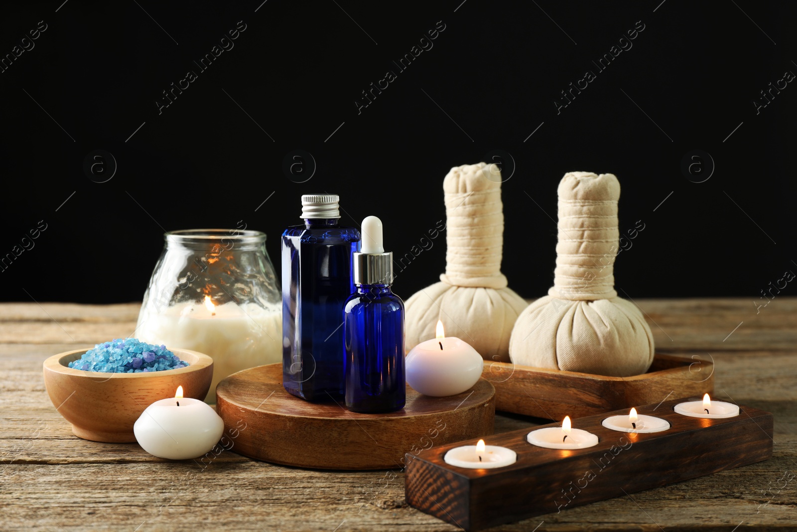 Photo of Spa composition. Cosmetic products, herbal bags, burning candles and sea salt on wooden table against black background