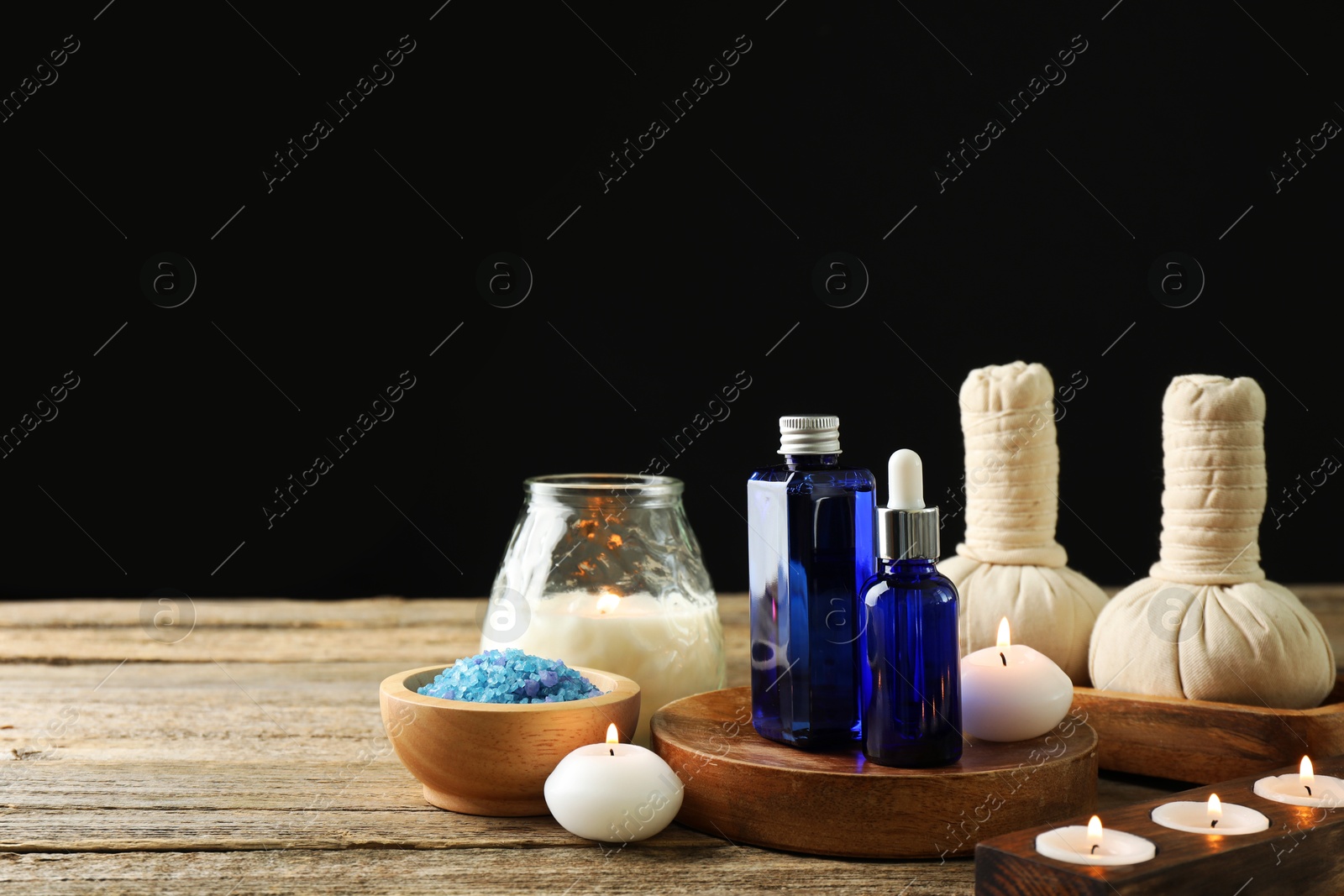 Photo of Spa composition. Cosmetic products, herbal bags, burning candles and sea salt on wooden table against black background, space for text