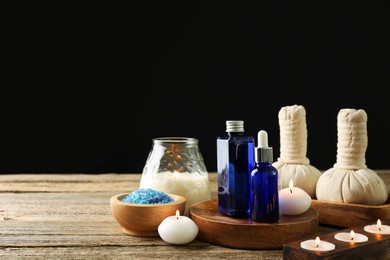 Photo of Spa composition. Cosmetic products, herbal bags, burning candles and sea salt on wooden table against black background, space for text