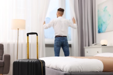 Guest opening curtains in stylish hotel room, focus on suitcase