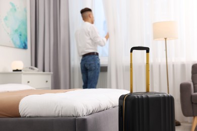 Guest opening curtains in stylish hotel room, focus on suitcase