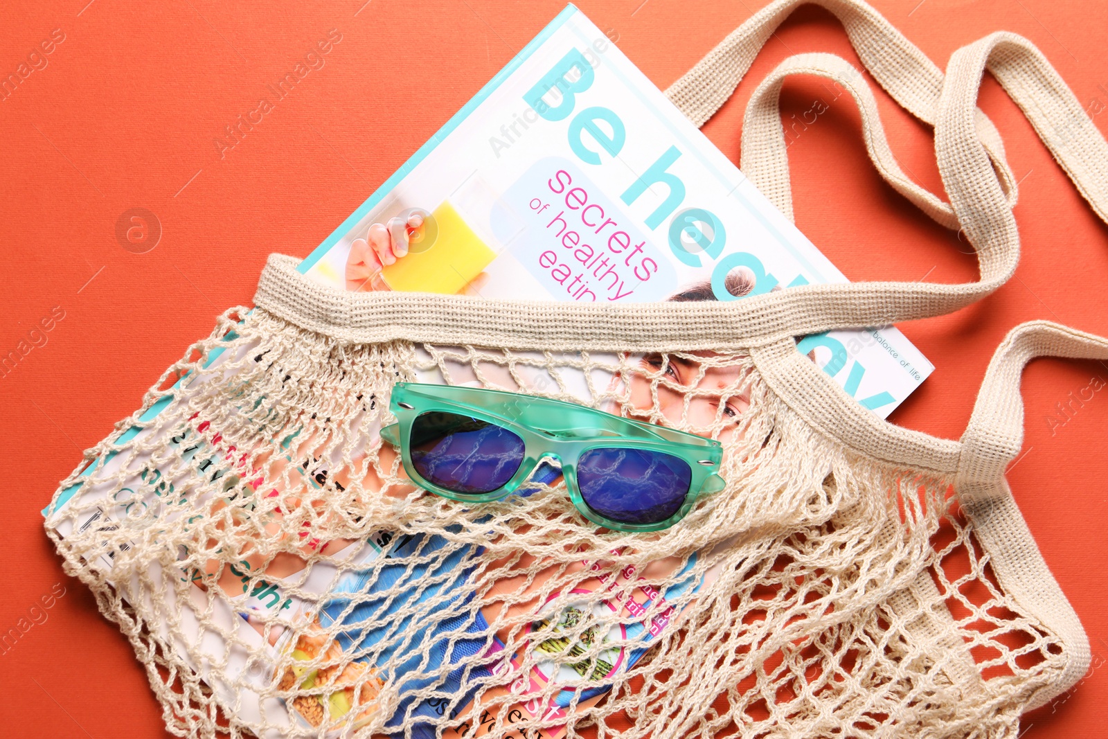 Photo of String bag, sunglasses and magazine on coral background, top view