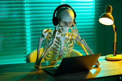 Photo of Waiting concept. Human skeleton sitting at wooden table with laptop and headphones indoors