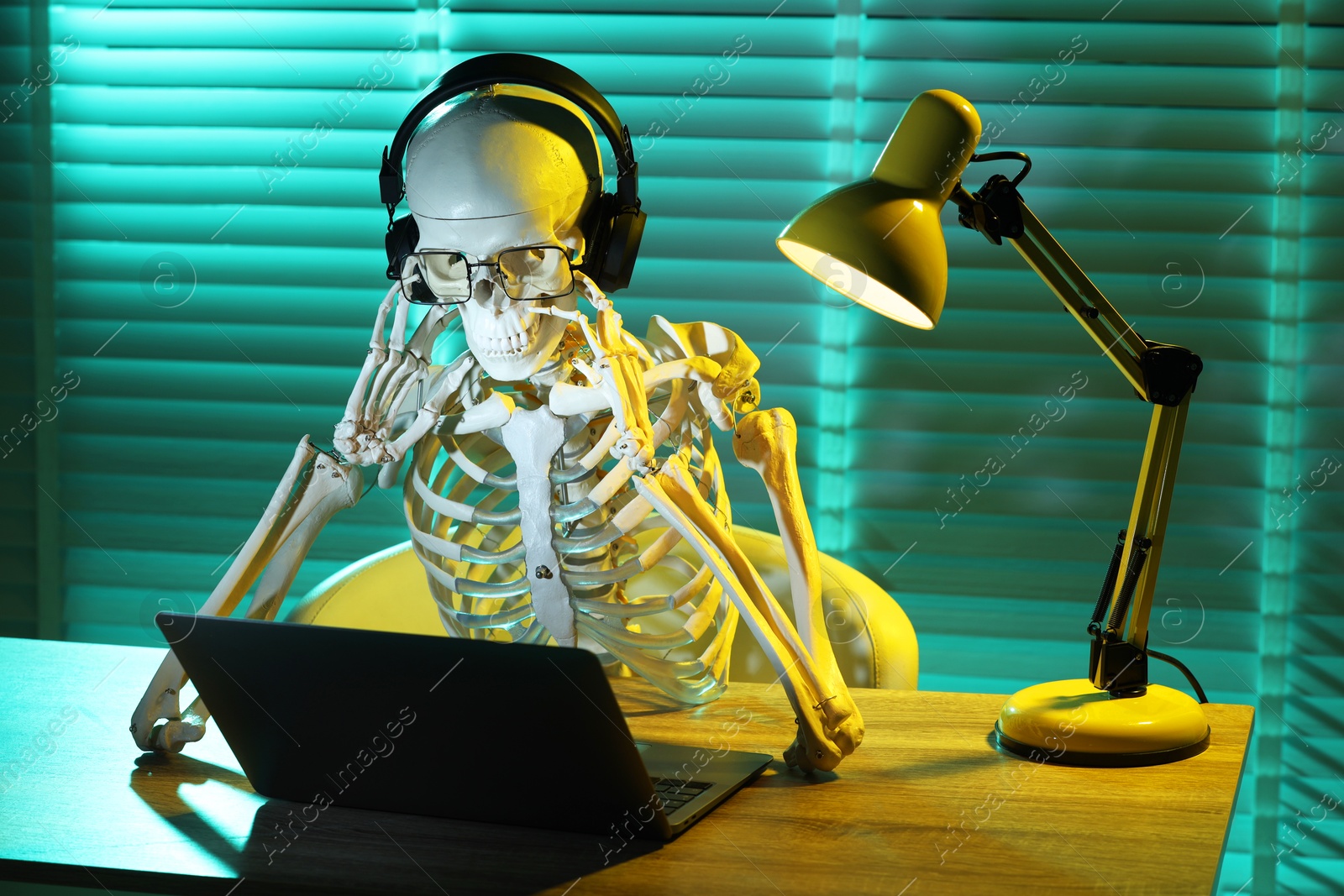 Photo of Waiting concept. Human skeleton sitting at wooden table with laptop and headphones indoors