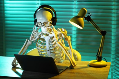Waiting concept. Human skeleton sitting at wooden table with laptop and headphones indoors