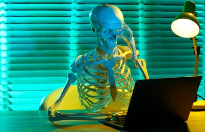 Photo of Waiting concept. Human skeleton sitting at wooden table with laptop indoors