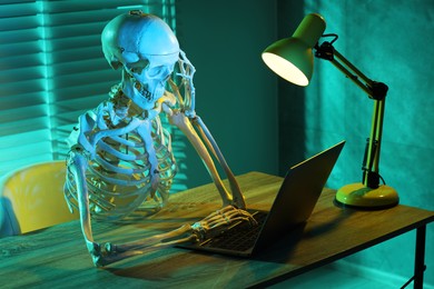 Photo of Waiting concept. Human skeleton sitting at wooden table with laptop indoors