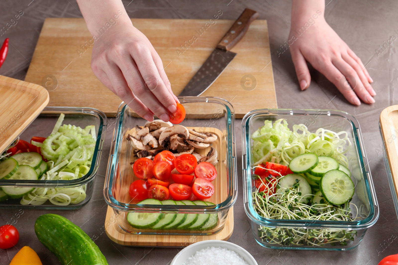 Photo of Healthy food. Woman putting piece of tomato into glass container at grey table, closeup