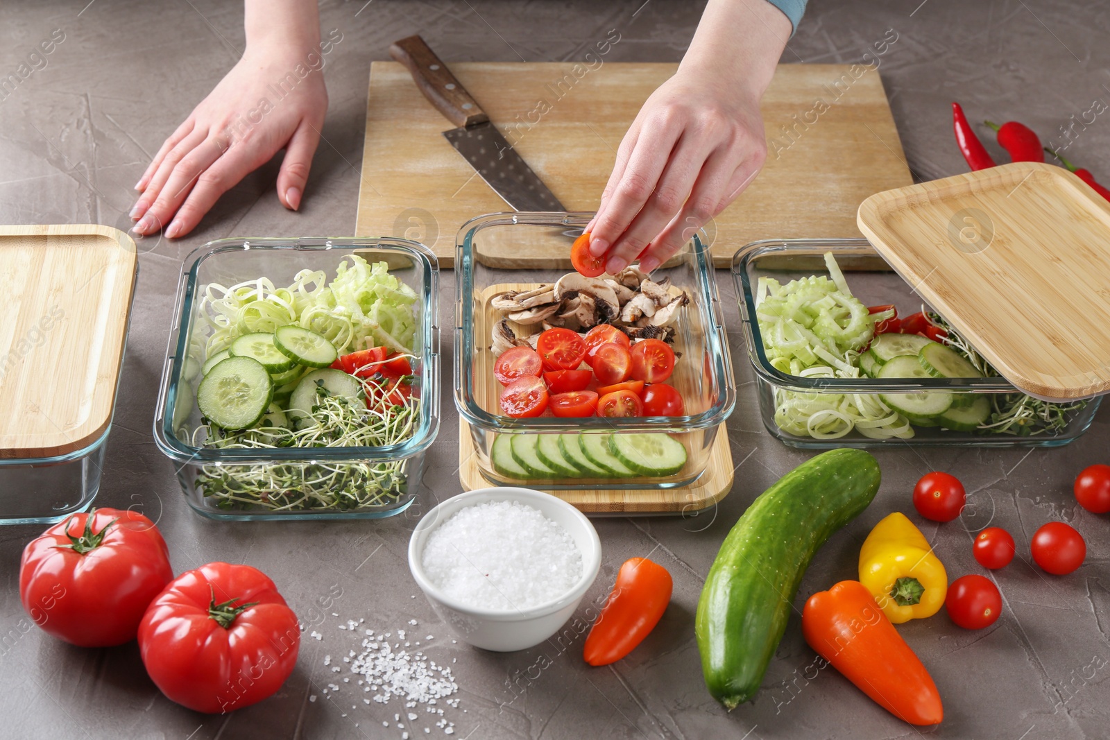 Photo of Healthy food. Woman putting piece of tomato into glass container at grey table, closeup