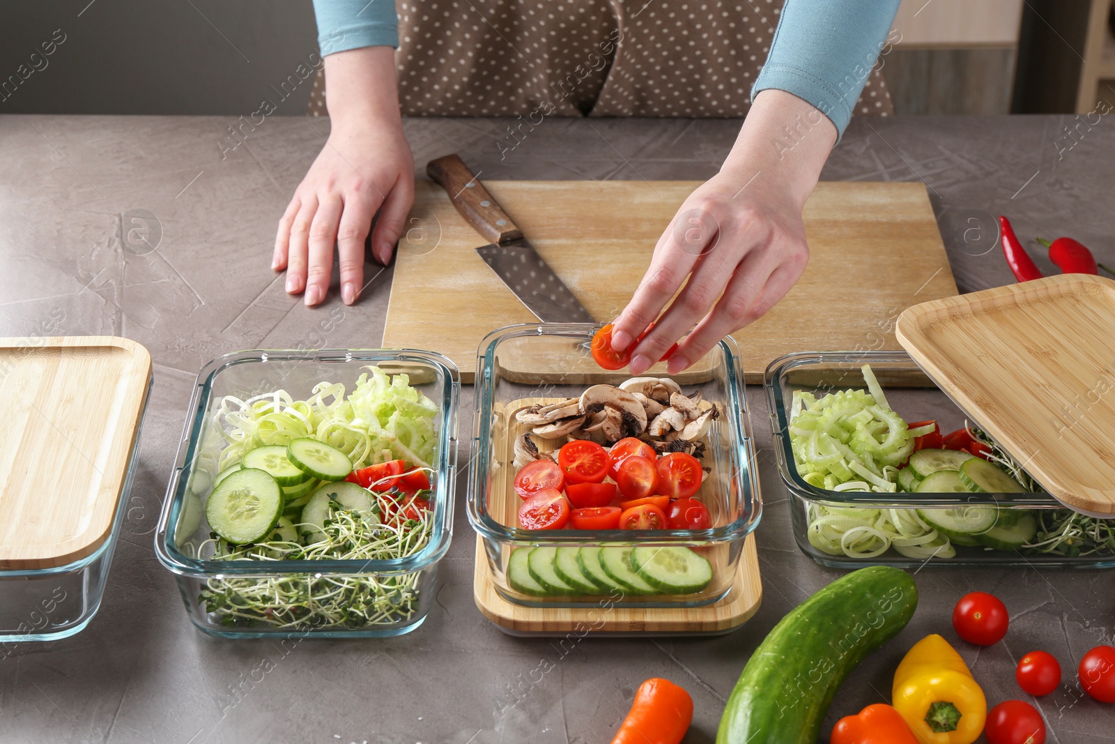 Photo of Healthy food. Woman putting piece of tomato into glass container at grey table in kitchen, closeup