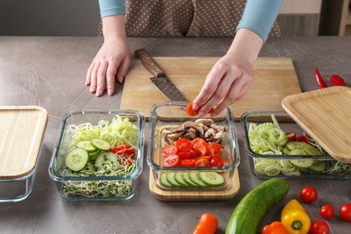 Healthy food. Woman putting piece of tomato into glass container at grey table in kitchen, closeup