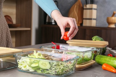 Photo of Healthy food. Woman putting piece of tomato into glass container at grey table in kitchen, closeup