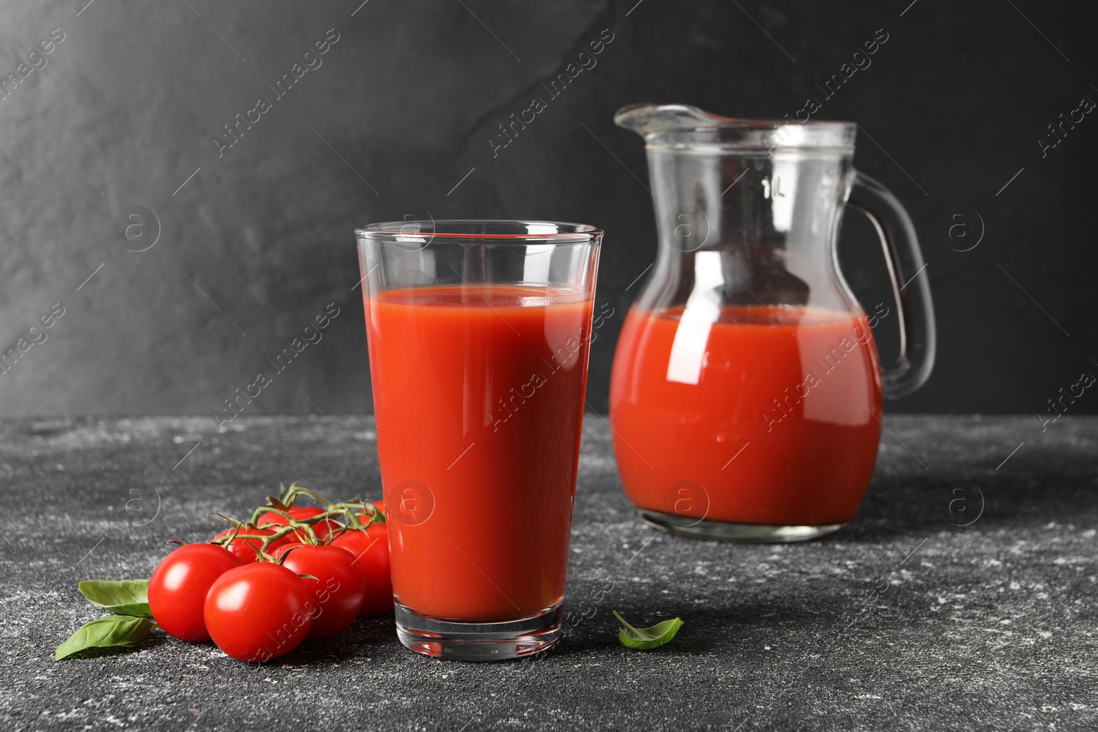 Photo of Tasty tomato juice in glass, jug, basil and fresh vegetables on grey table