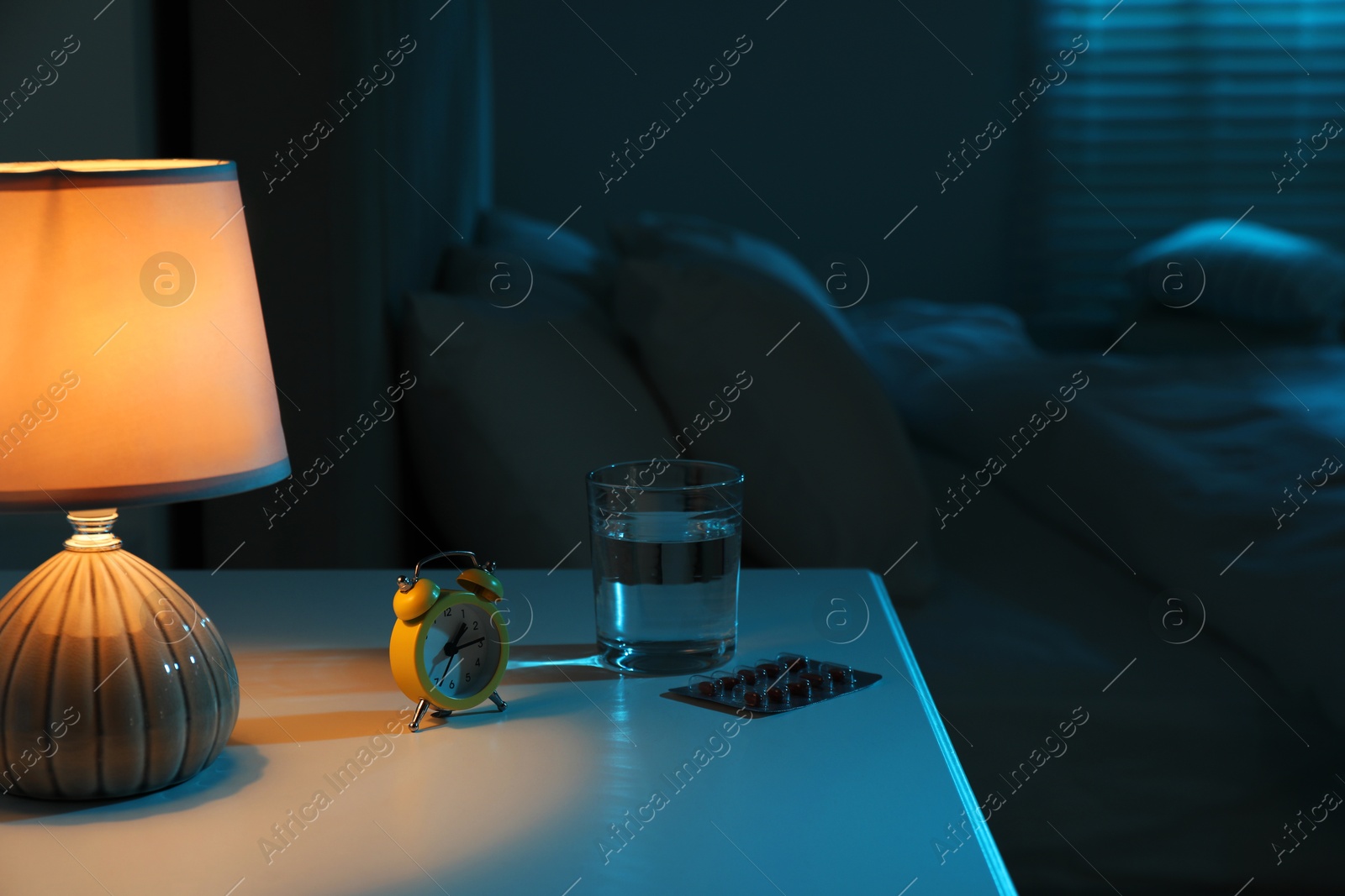 Photo of Insomnia treatment. Glass of water, pills and alarm clock on bedside table in bedroom at night