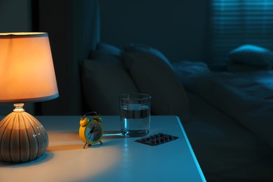 Photo of Insomnia treatment. Glass of water, pills and alarm clock on bedside table in bedroom at night