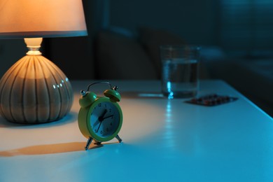 Insomnia treatment. Glass of water, pills and alarm clock on bedside table in bedroom at night, selective focus