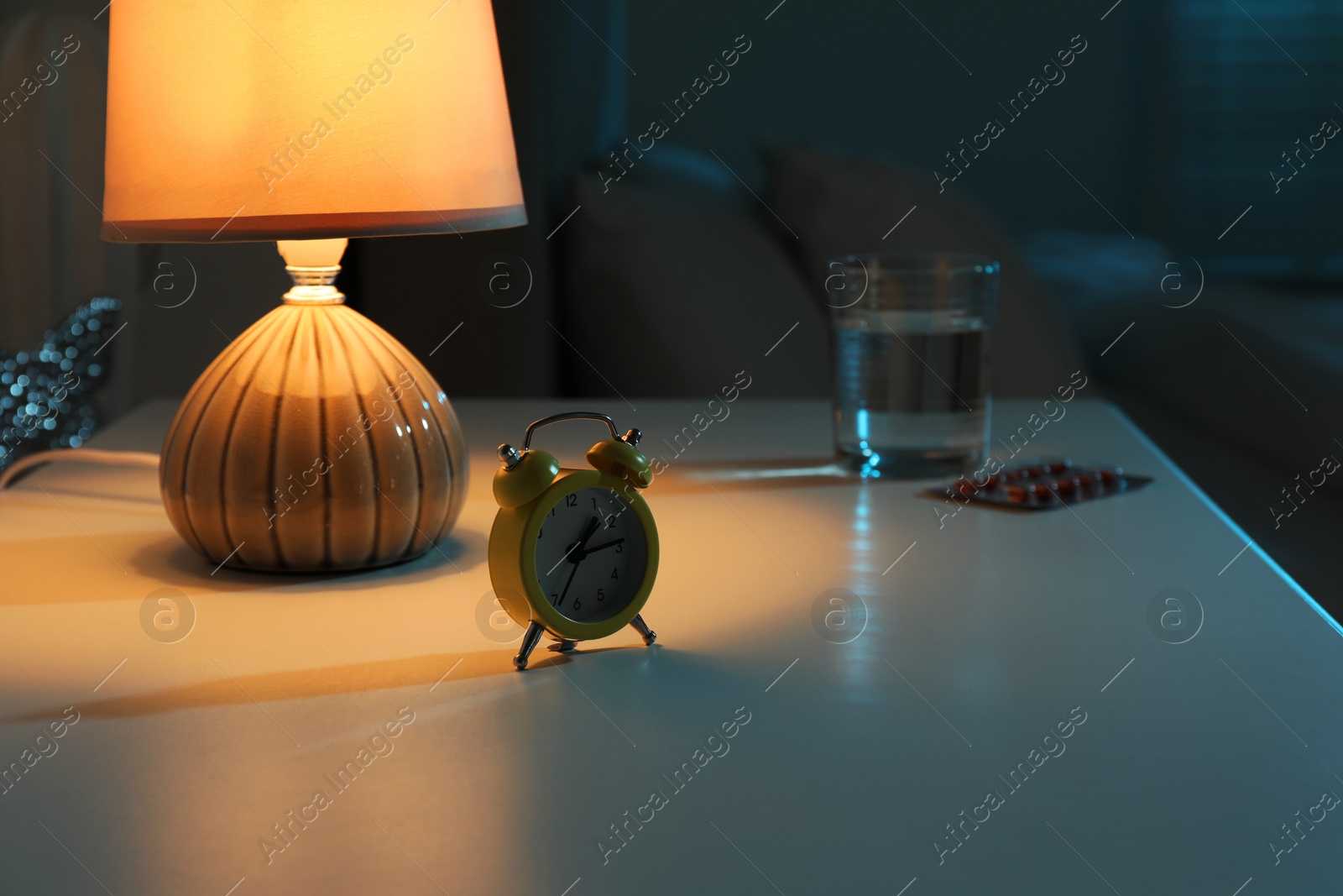 Photo of Insomnia treatment. Glass of water, pills and alarm clock on bedside table in bedroom at night, selective focus