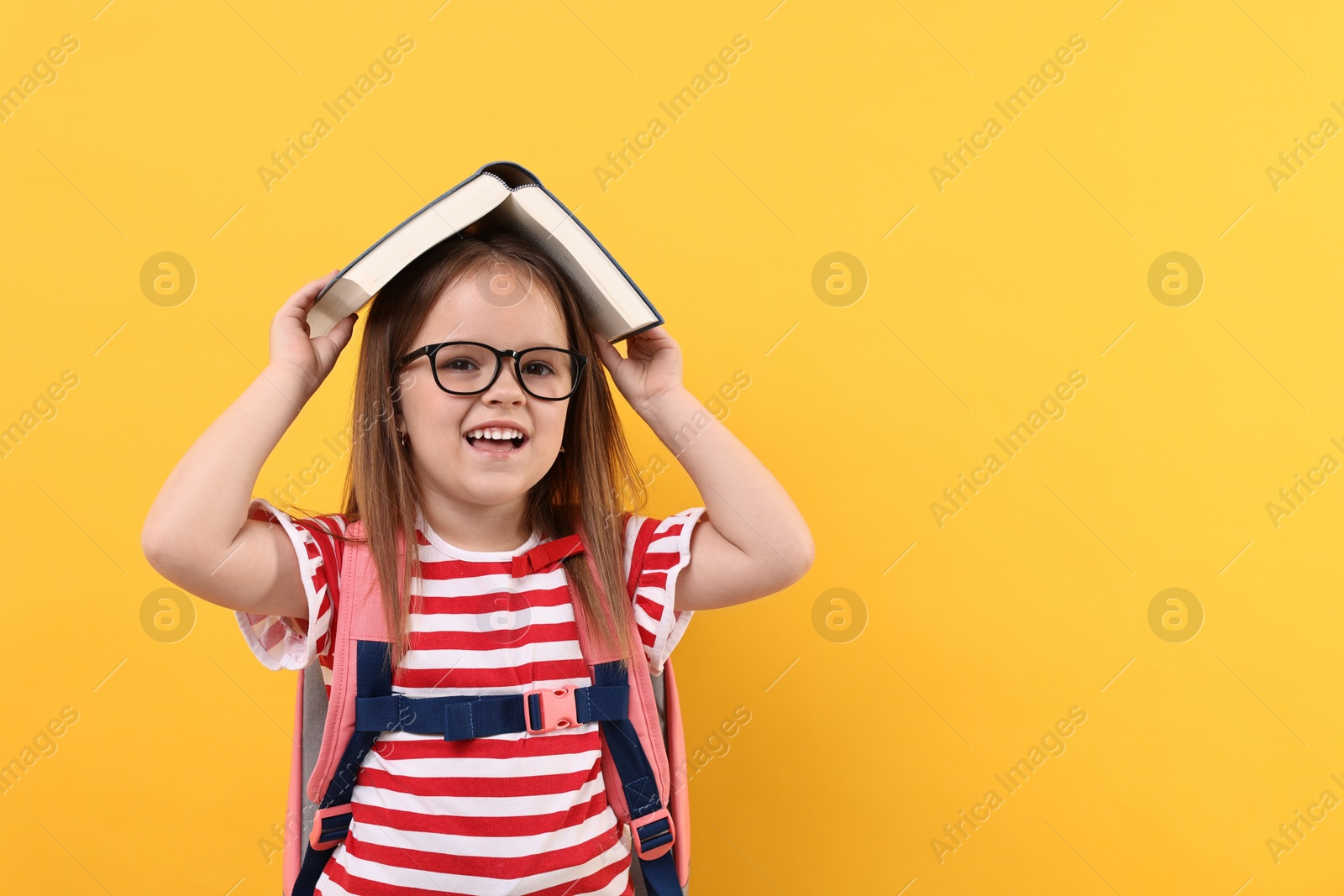 Photo of Cute little girl in glasses with open book and backpack against orange background. Space for text