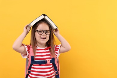 Photo of Cute little girl in glasses with open book and backpack against orange background. Space for text